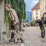 Paved street, a wood sculpture of a man and dog, with a boy riding on one wheel a mountain bike.