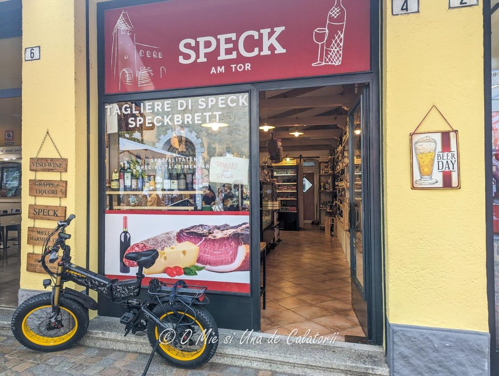 the entrance of a small shop, selling food and drinks, and also making panini and speck, with an electric bicycle left out front
