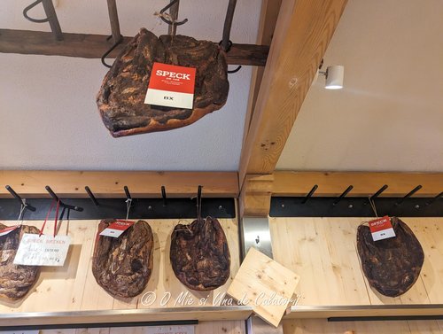 a interior photo of the shop, with multiple pieces of speck (a local meat) hanged on hooks for sale.