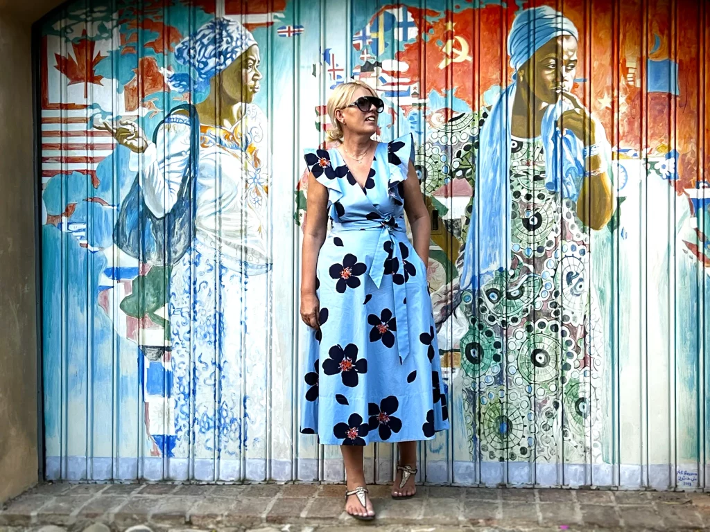 A woman posing in front of a garage door depicting two persons of colour in traditional clothing and a world map made of flags