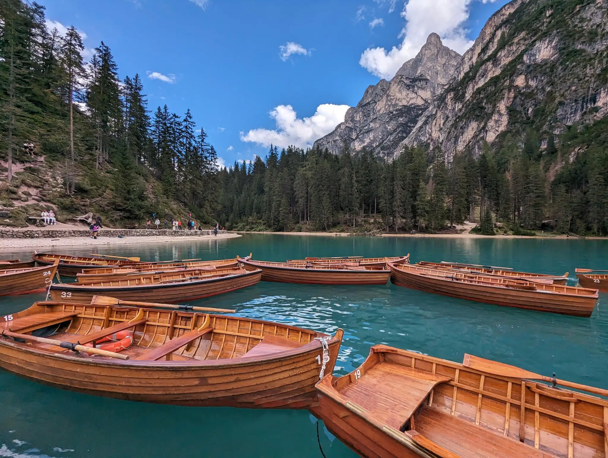 A clear blue lake with rowing boats in the close plan, people along the shore and mountains in the background