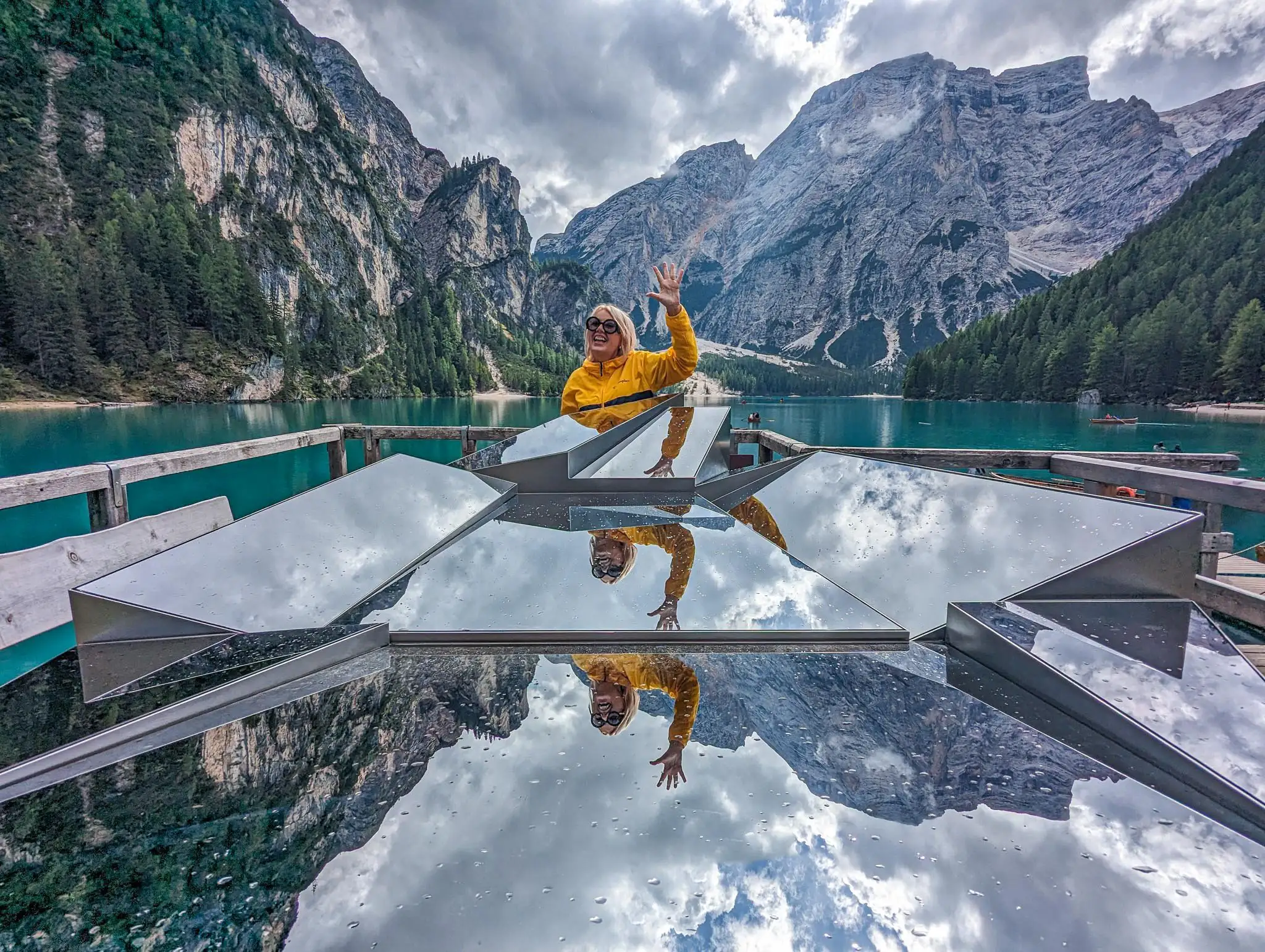 a person waving her hand in front of a mirrored abstract art structure with the lane behind and mountains