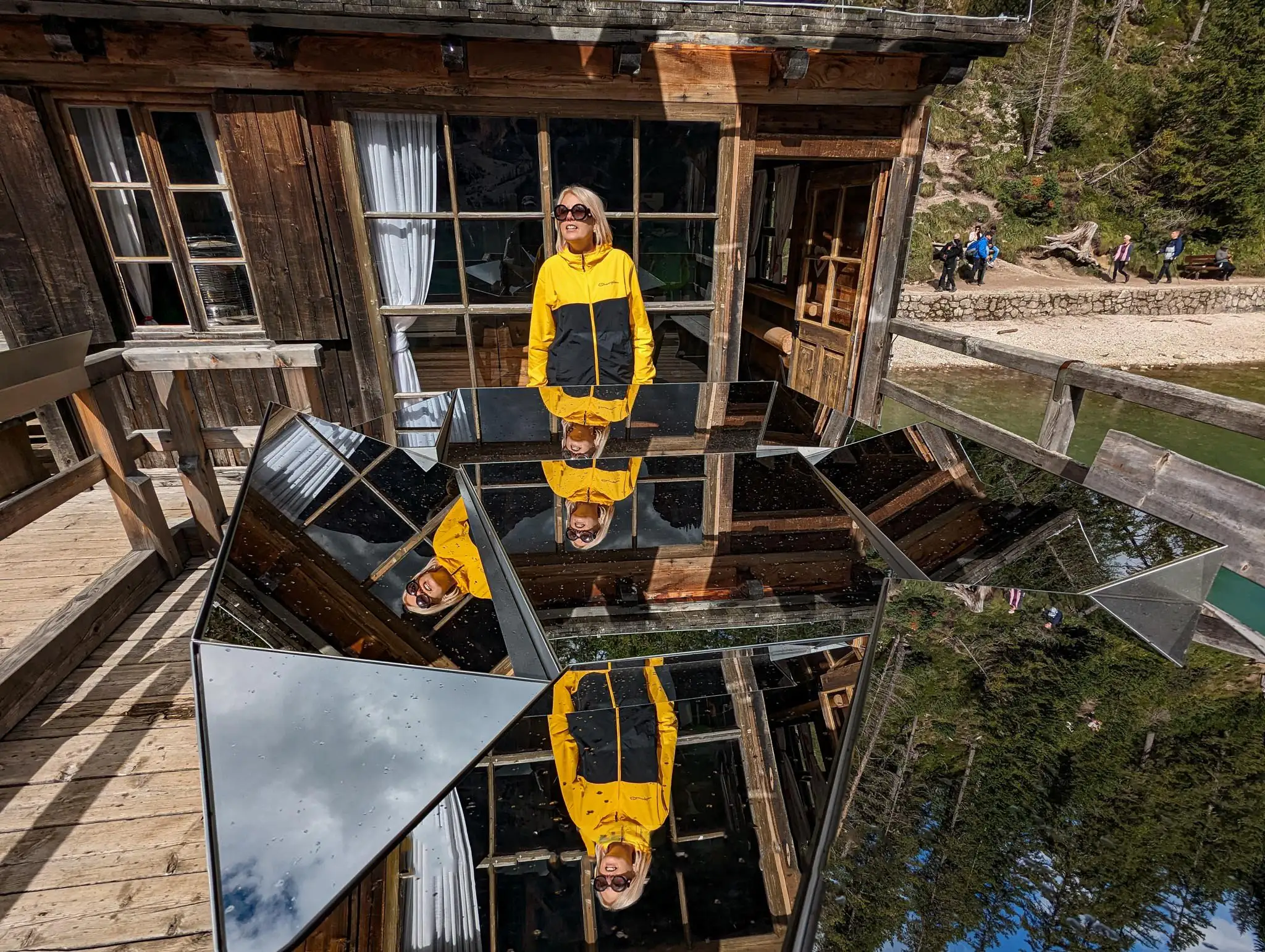 a person waving her hand in front of a mirrored abstract art structure with a cabin in the background and trees