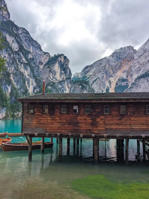 a wood cabin on wood pylons on the lake, with row boats tied to it, mountains in the background