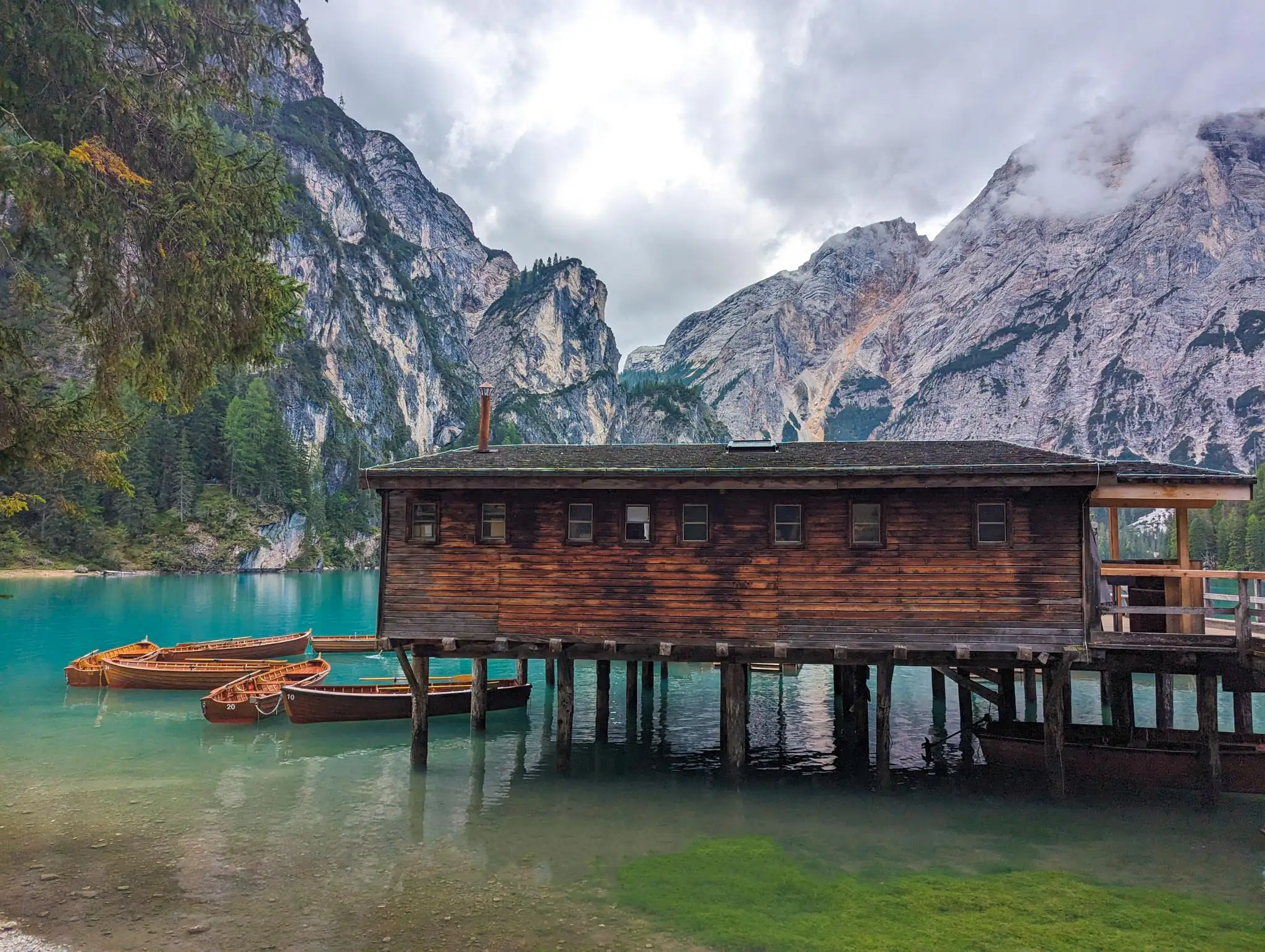 a wood cabin on wood pylons on the lake, with row boats tied to it, mountains in the background
