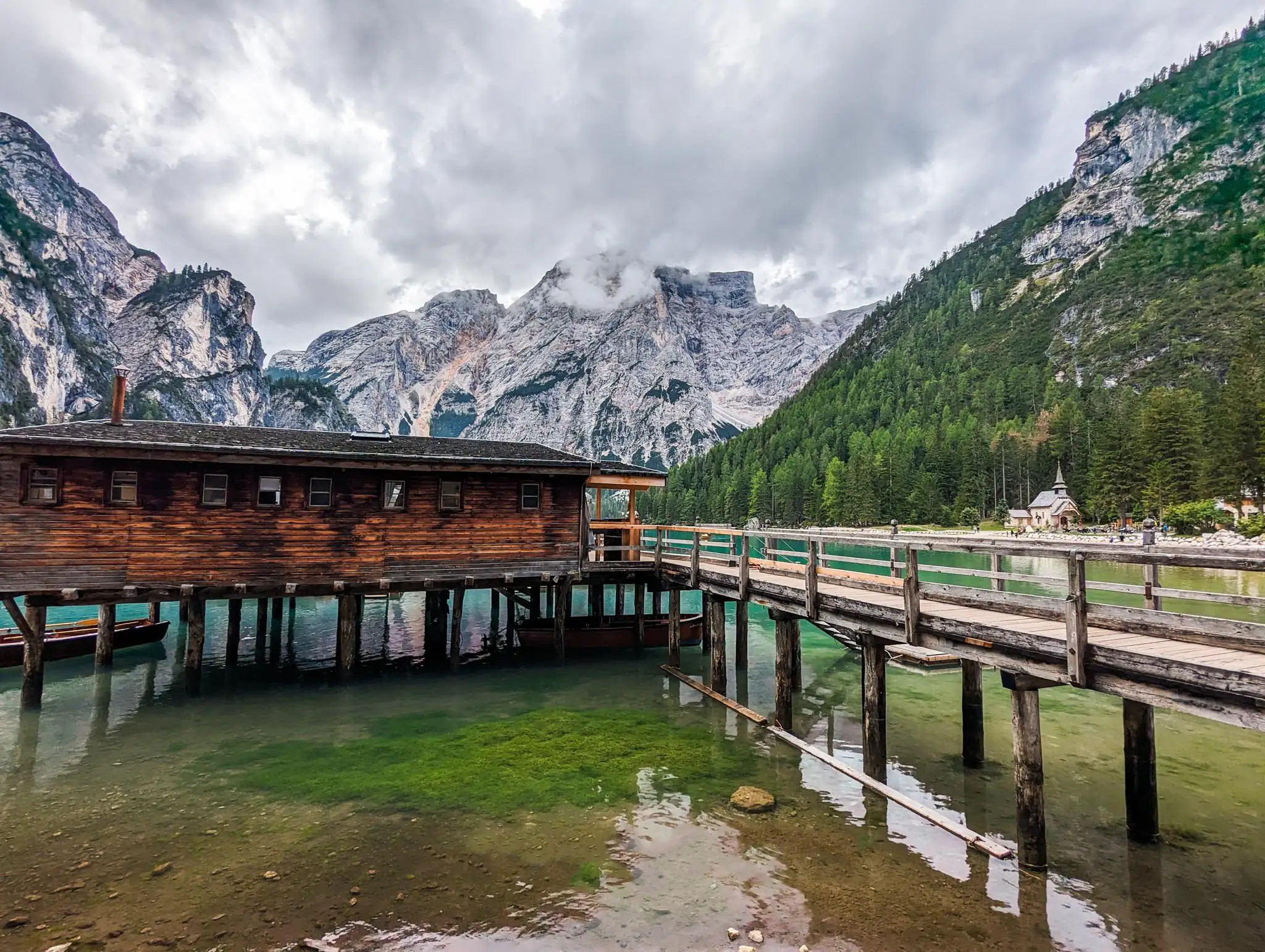 a different angle of the wood cabin on wood pylons on the lake, with row boats tied to it, mountains in the background and a small church