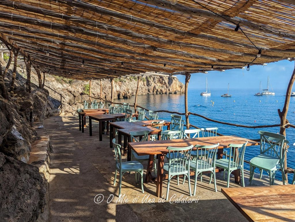 A beautiful shot of a sea side terrace, with multiple tables and chairs, covered with a shade and multiple boats in the far background on a blue sea