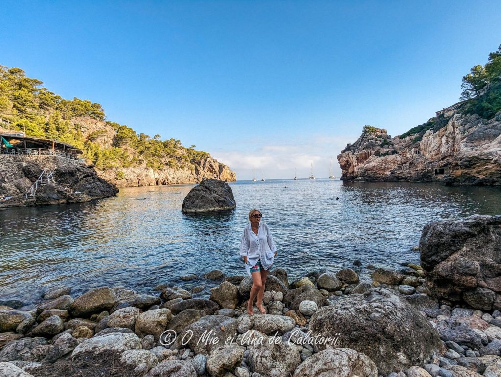 a shot of a person posing for the camera in front of the sea, with a restaurant on the far left, boats on the middle, and rocks on the right