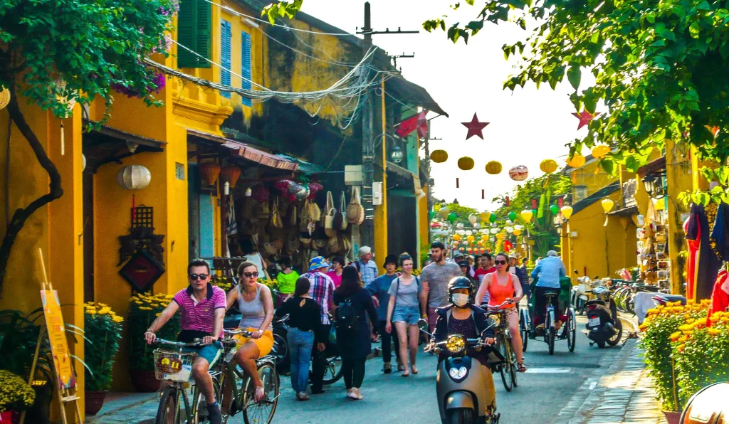 a busy street with many people on bicycles walking, coloured lanterns all over the place and shops