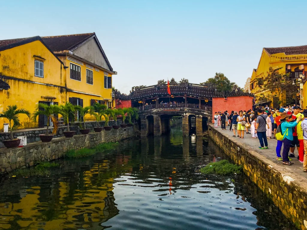 A shot of a bridge over a patch of water with many tourists along the right shore and pots with palm trees on the left with yellow houses in the background.