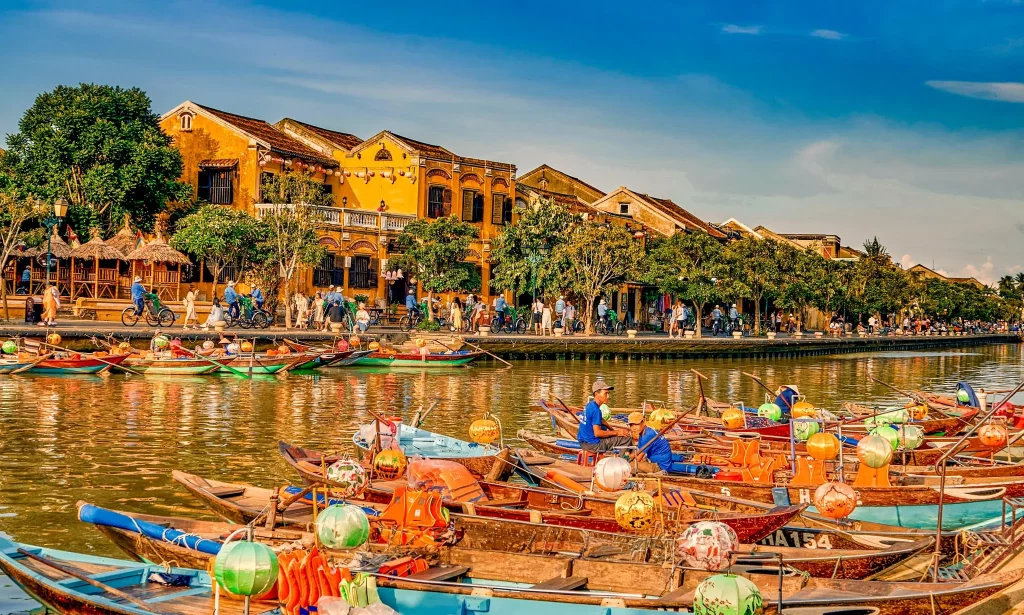 Multiple traditional fishing boats along the close shore with tourists on the opposite shore, many trees and yellow houses