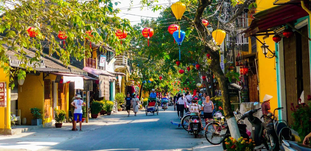 a street with tourists, green trees on both sides and traditional houses and hanging lanterns