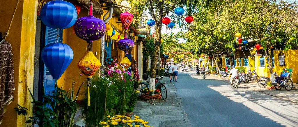 a street with tourists, green trees on both sides and traditional houses and hanging lanterns