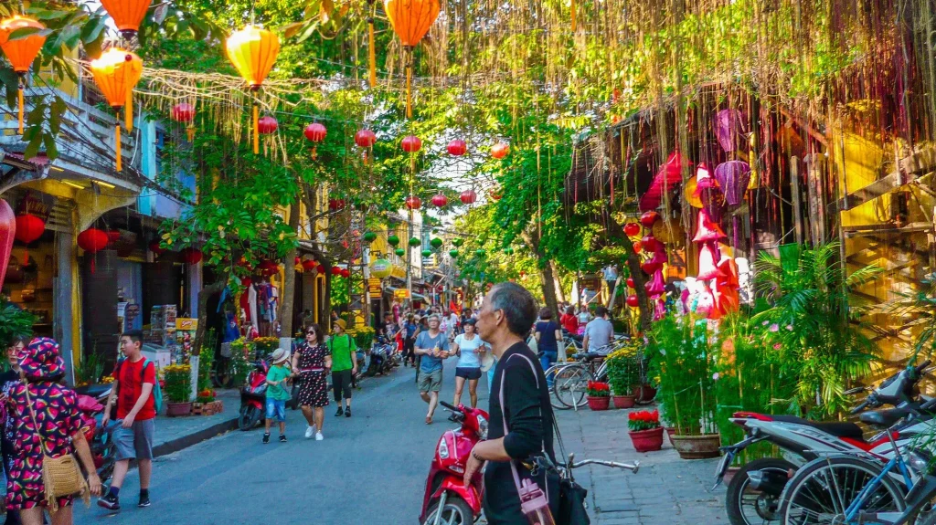a street with tourists, green trees on both sides and traditional houses and hanging lanterns