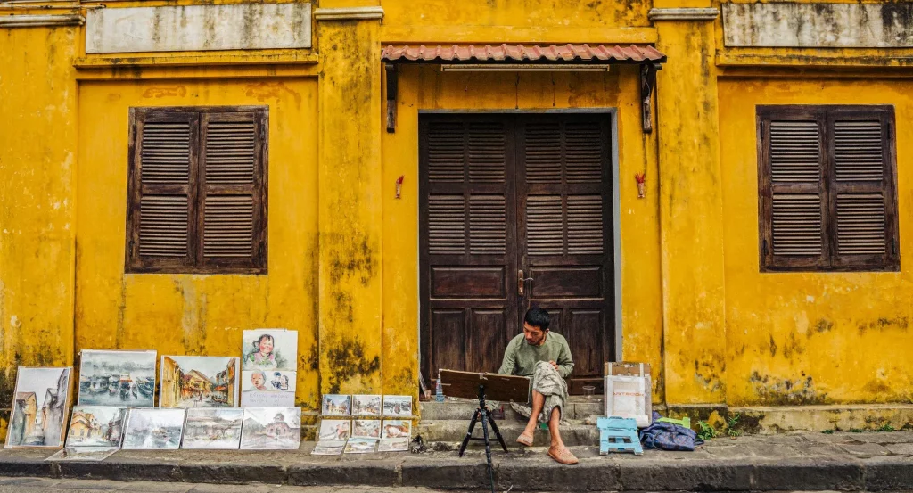 A street painter doing works of art and presenting some for sale sitting in front of a yellow house