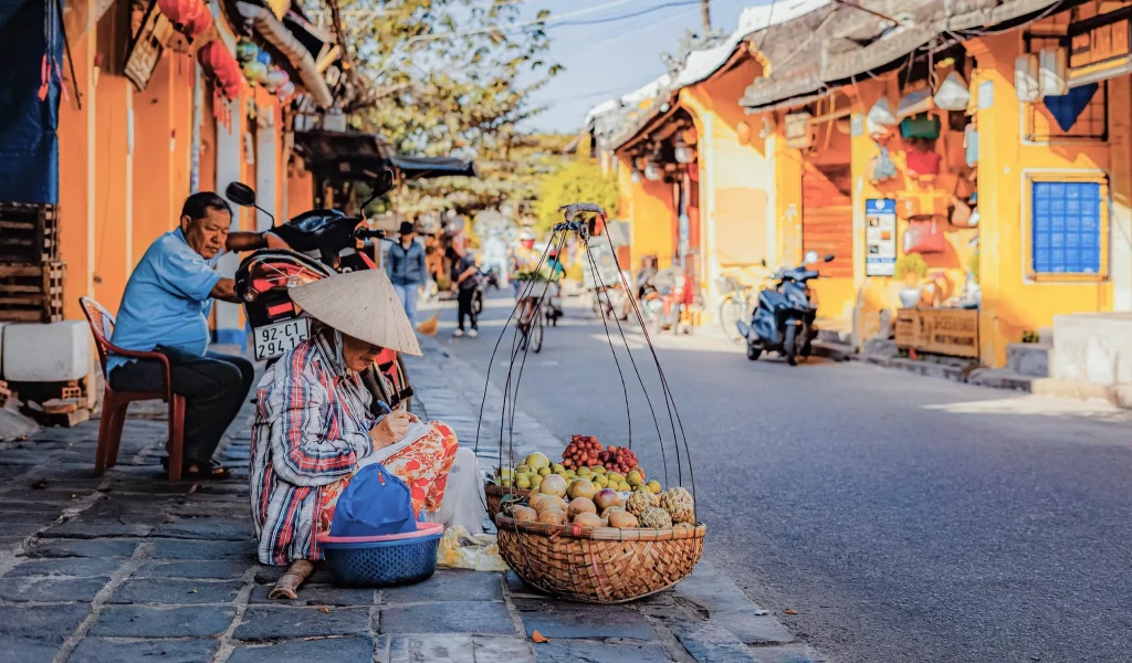 A street from Hoi An with a lady selling fruits in the street, some traditional baskets, you can see a few shops in the background