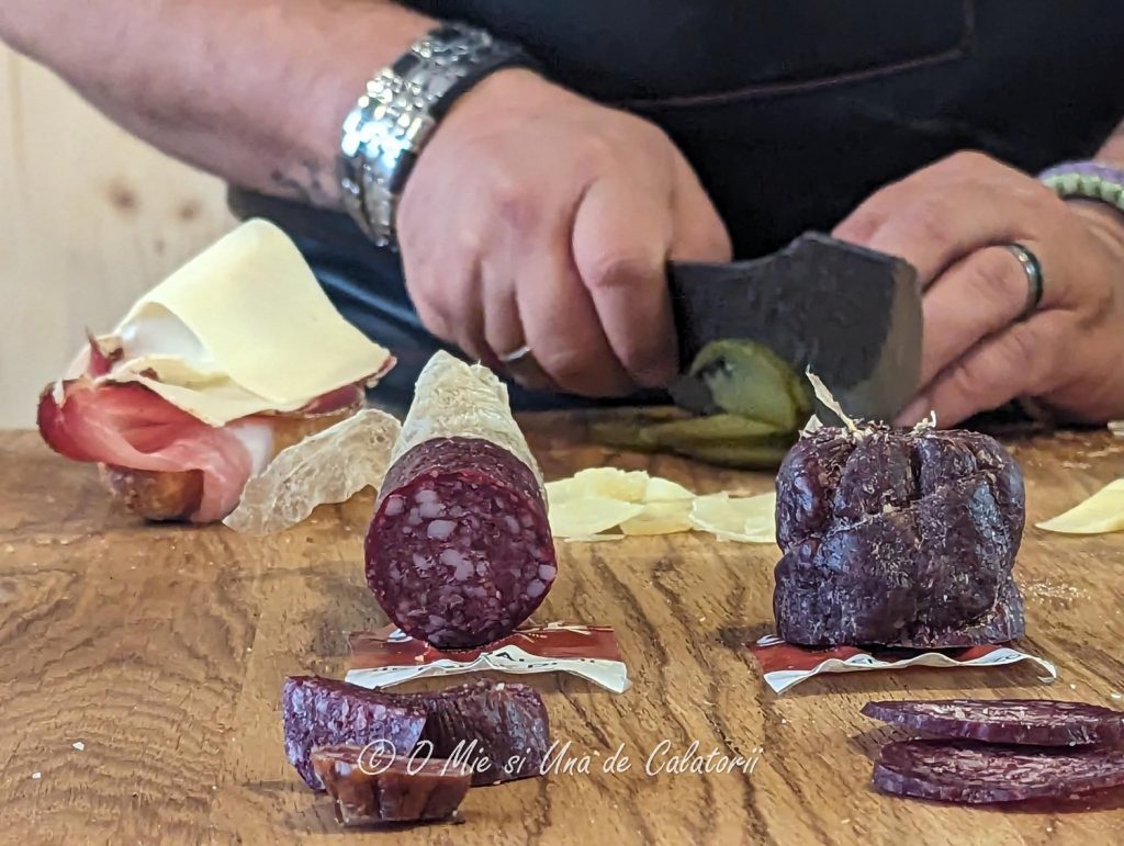 a close up of a knife chopping up some pickles with some meat products also showing, as the shop seller is preparing a sandwich.